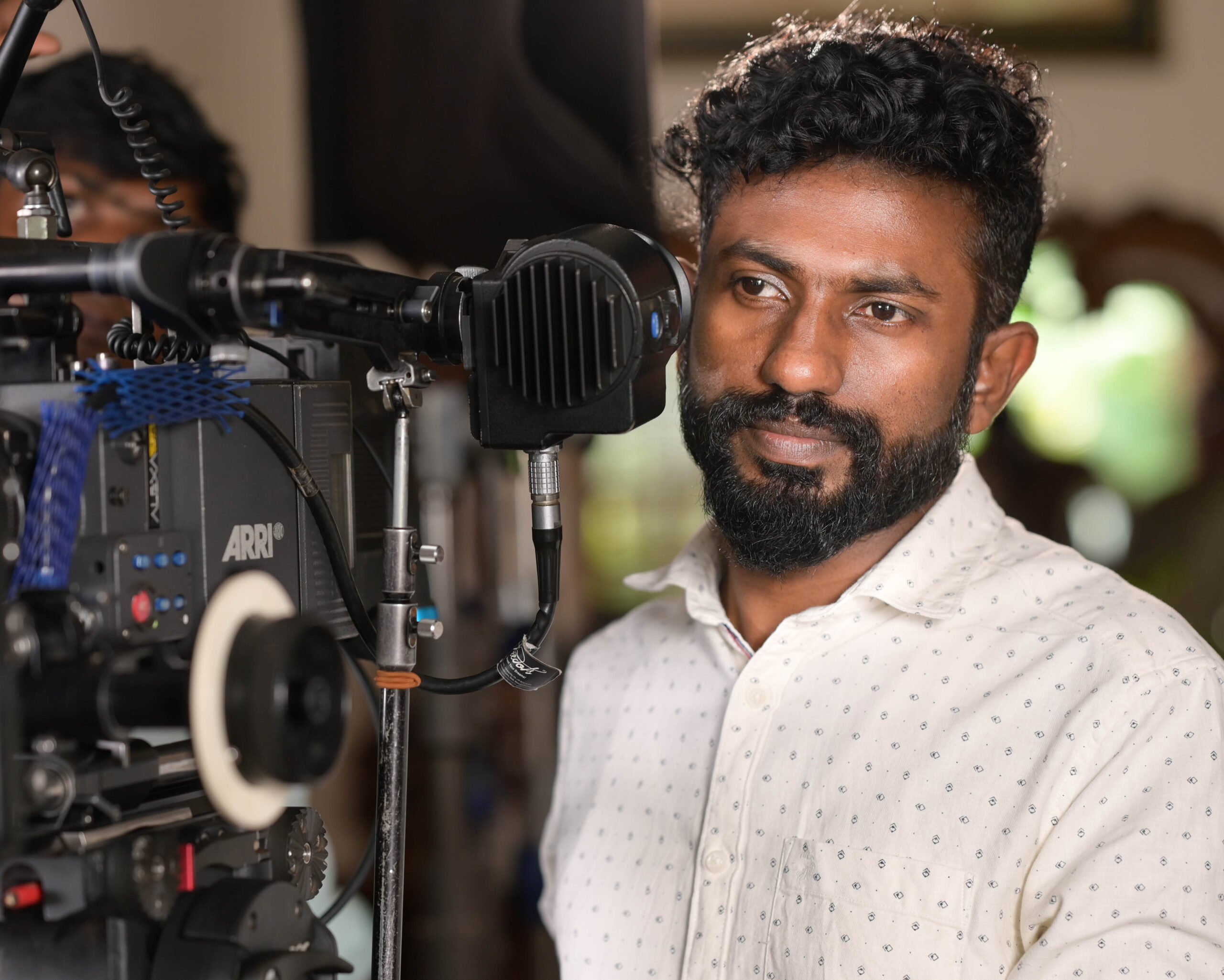 Professional cinematographer standing beside a fully rigged ARRI cinema camera on a film set, focusing intently on the shot while wearing a light-colored patterned shirt. The camera is mounted on a tripod with matte box, follow focus, cables, and monitoring accessories visible, highlighting a behind-the-scenes filmmaking environment with soft indoor lighting and a blurred background suggesting crew activity.